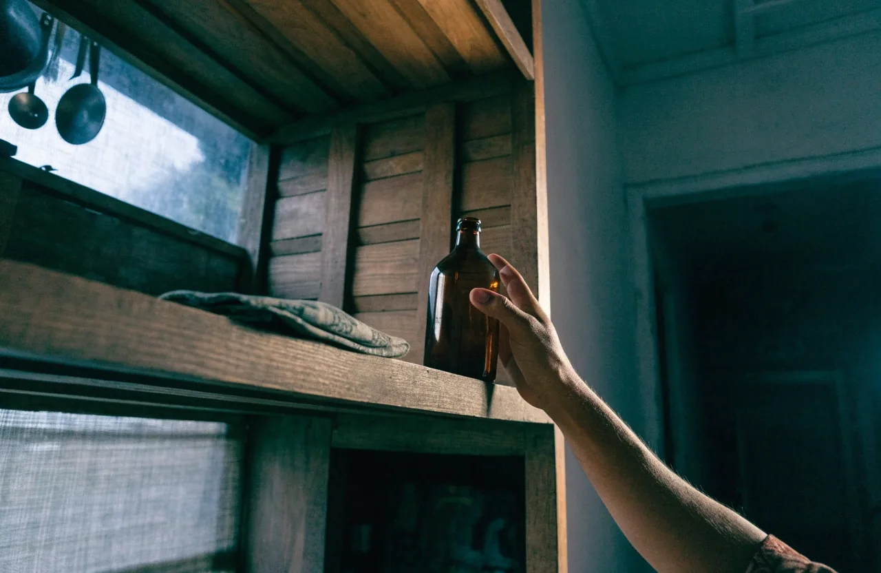 Unlabeled matte bottles stored upright on a cool shaded shelf beside a damp window edge